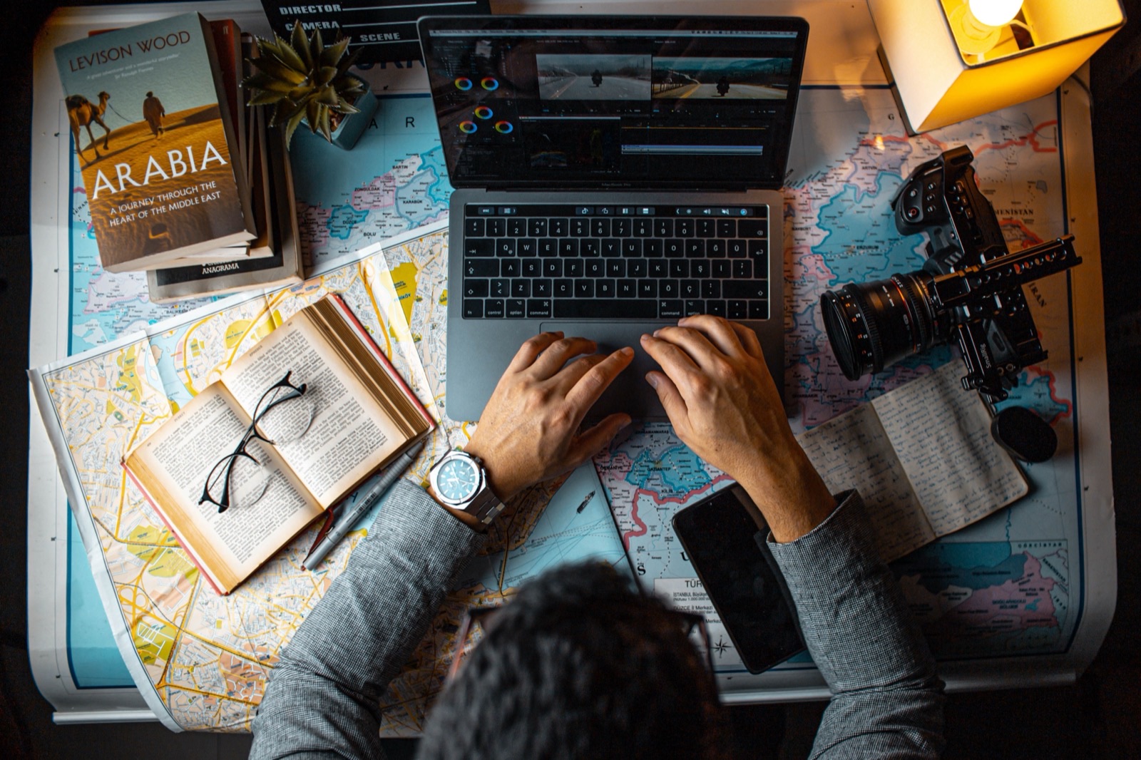 Travel content creator planning a marketing strategy — editing destination footage on a laptop surrounded by maps, a camera and travel notes